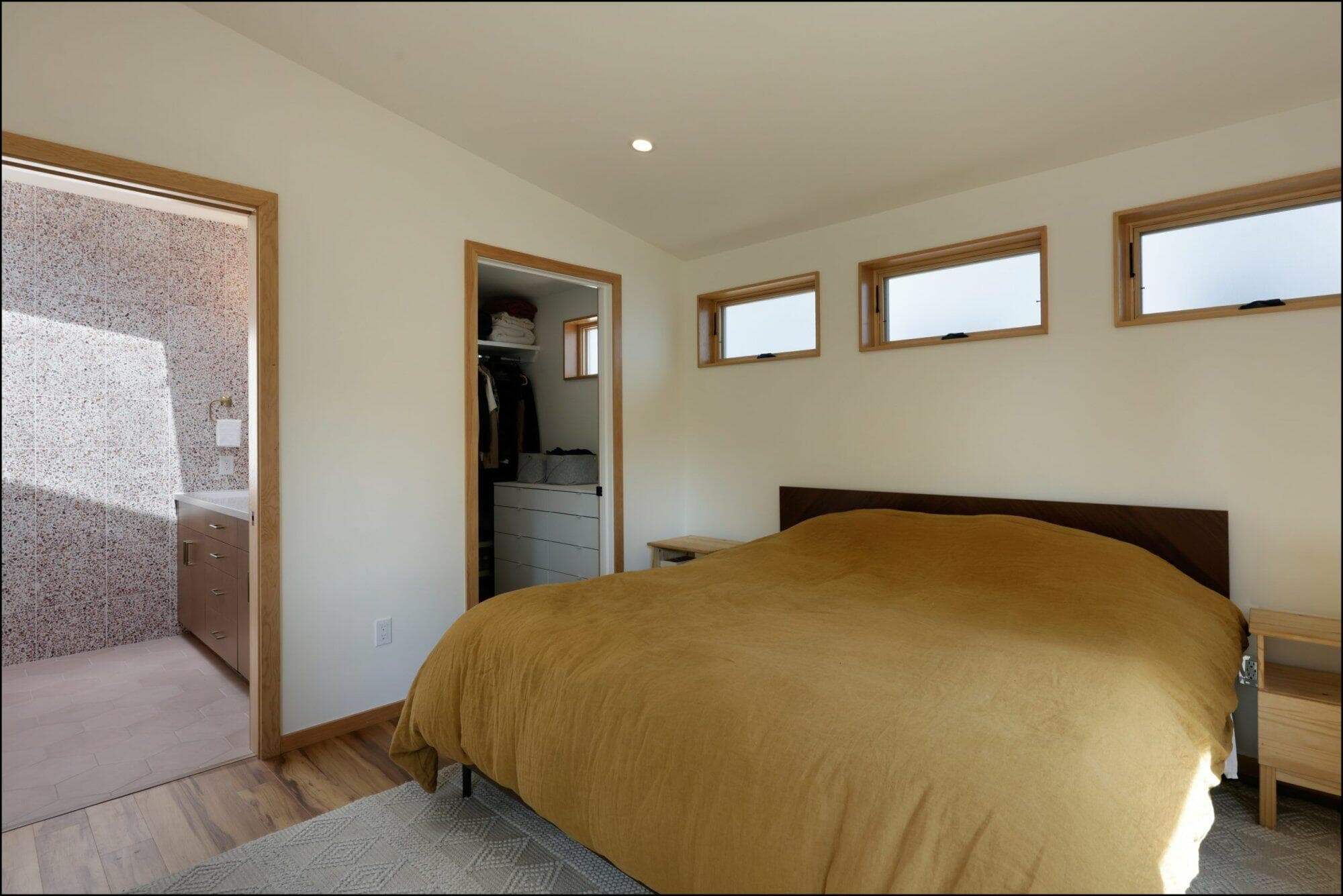 Modern bedroom with a brown bedspread, showcasing an entrance to a bathroom and closet, featuring natural light from multiple windows, highlighting ReCraft Home Remodeling's interior design work.