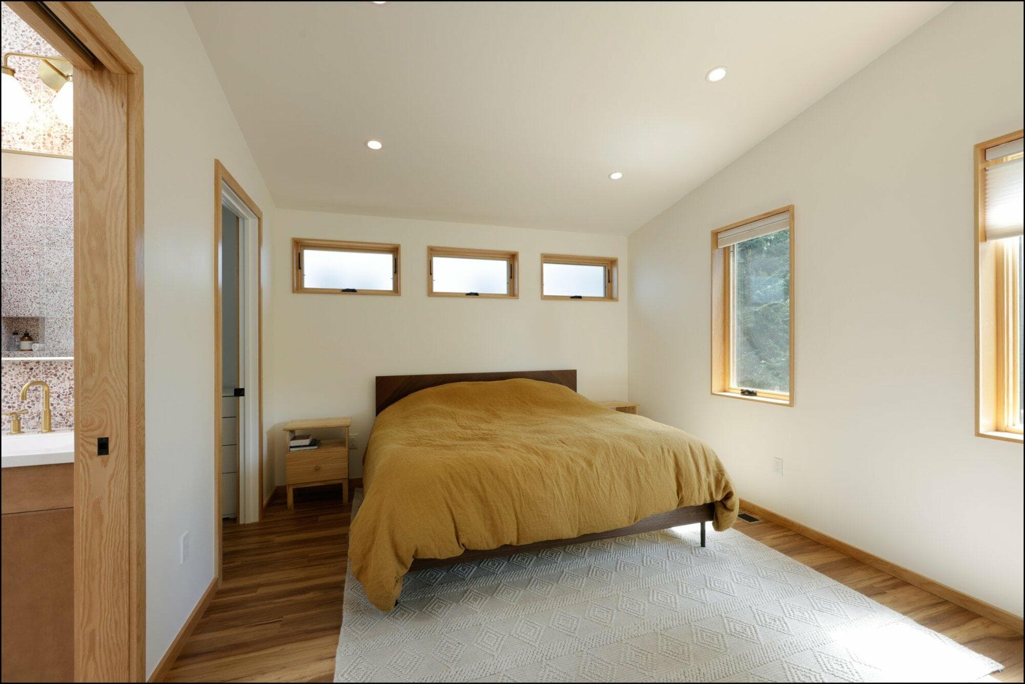Modern bedroom interior showcasing a warm-toned bedspread, natural wood accents, and ample natural light from multiple windows, reflecting ReCraft Home Remodeling's design aesthetic in Portland.