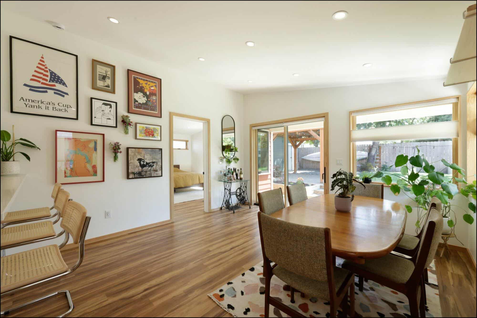 Dining room with wooden flooring, round table, and chairs, featuring framed artwork on walls and plants, leading to a bright entryway towards a bedroom, showcasing ReCraft Home Remodeling's design aesthetic.