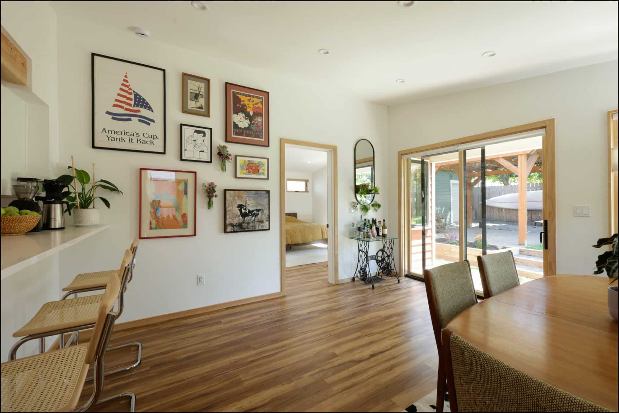 A well-lit dining area with a wooden table and four chairs. The room has a wall adorned with framed pictures, a sliding glass door, and a doorway leading to a bedroom.