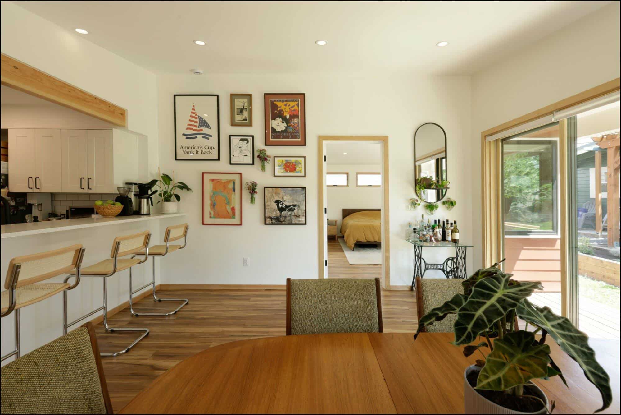 Modern dining area featuring a round wooden table, contemporary artwork on the walls, bar stools at a kitchen counter, and a view of a bedroom through an open doorway, showcasing ReCraft Home Remodeling's design aesthetic.