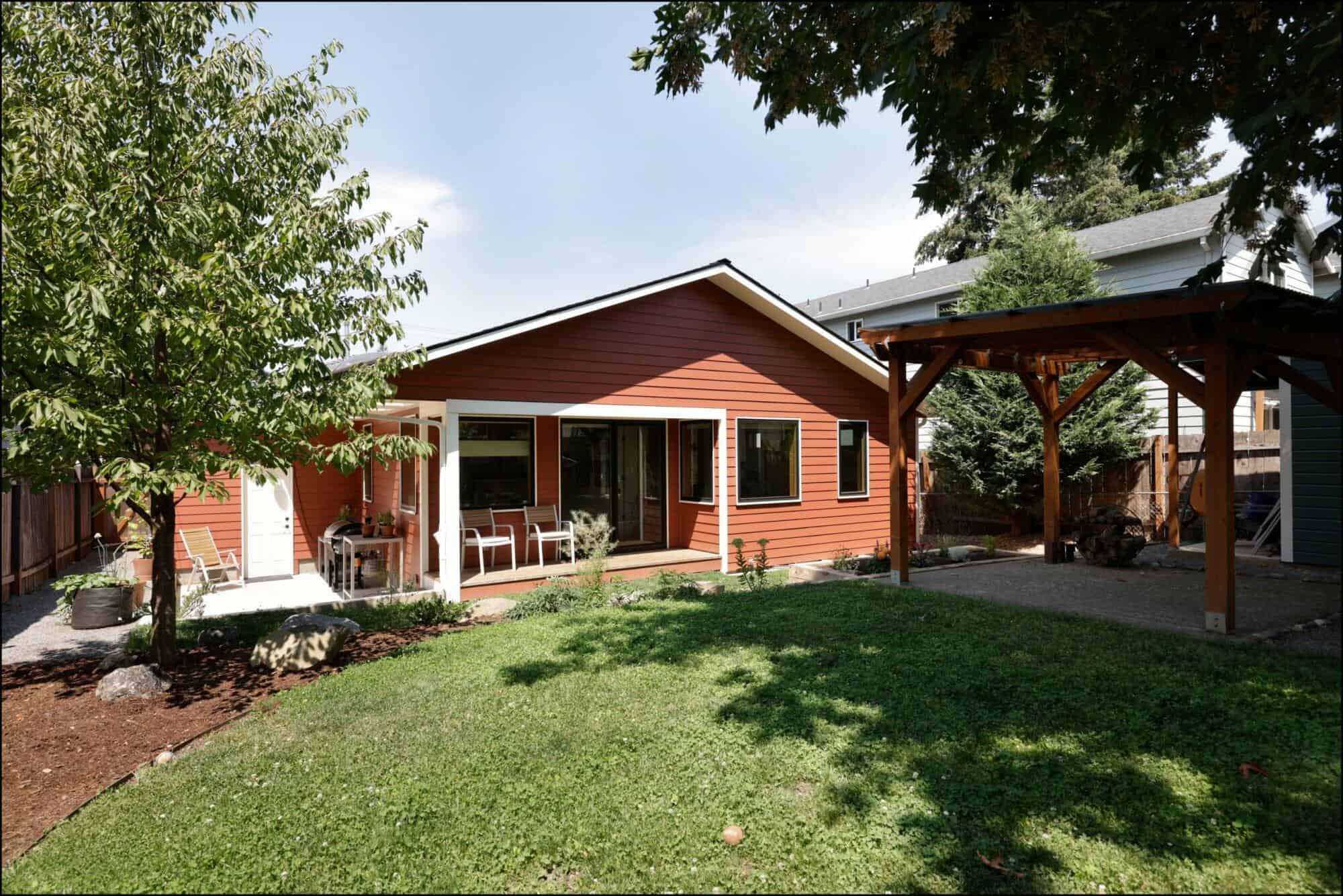 A charming red one-story home in Portland features a large glass door, a patio with chairs, and a well-maintained lawn. Its architectural highlights include a backyard with a tree and a wooden pergola, showcasing popular home styles of the region.