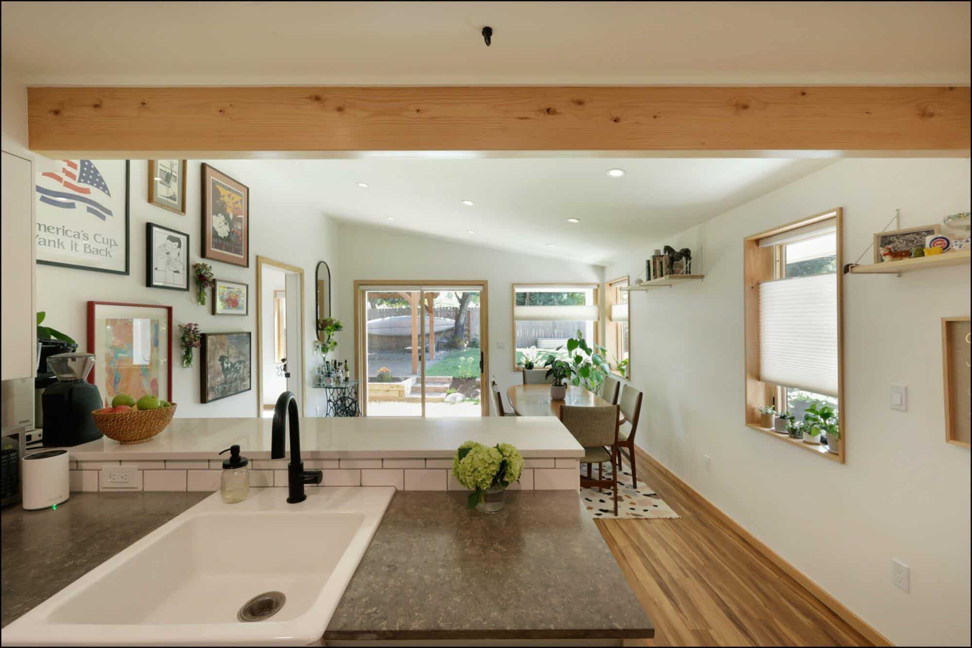 A modern kitchen with a sink and faucet, stone countertop, and light wood flooring, leading into a bright dining area with windows, framed artworks, and indoor plants.