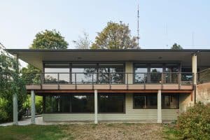 This Portland two-story modern house features large windows, a flat roof, and an upper-level balcony, seamlessly blending architectural elegance with its lush surroundings of trees and grass.