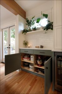 Open kitchen cabinet with appliances, a shelf above with potted plants, a mug, and a candle. White subway tile backsplash and French doors in the background.