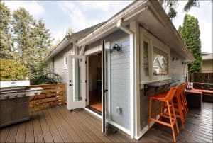 A modern outdoor deck with a grill, bar stools, and a small house exterior. The deck has dark wood flooring and is surrounded by trees and a fence.