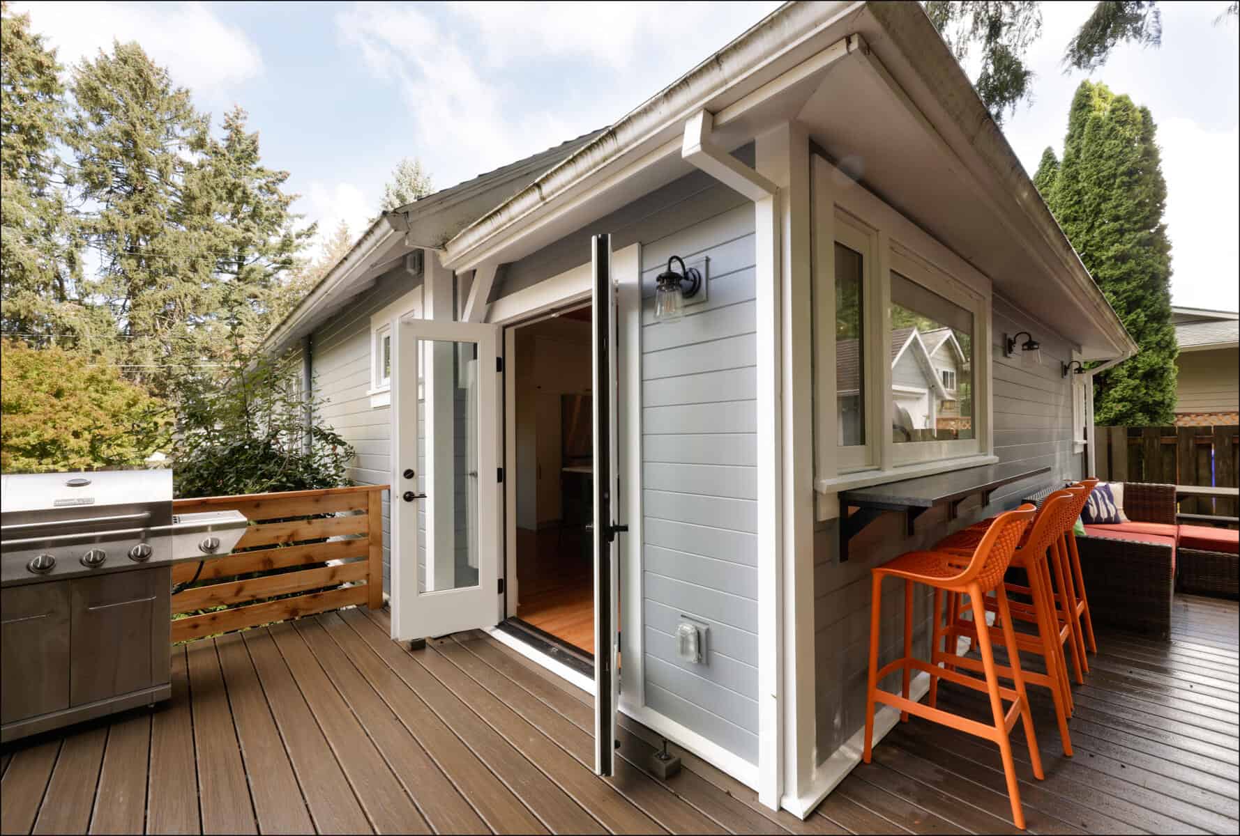 A modern outdoor deck with a grill, bar stools, and a small house exterior. The deck has dark wood flooring and is surrounded by trees and a fence. A modern outdoor deck with a grill, bar stools, and a small house exterior. The deck has dark wood flooring and is surrounded by trees and a fence.