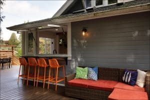 Outdoor patio with wicker sofa, colorful cushions, and orange bar stools at a counter window. Gray house siding and a single outdoor light above the seating area.
