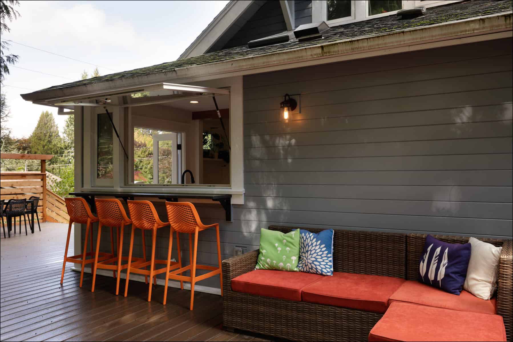 Outdoor patio with wicker sofa, colorful cushions, and orange bar stools at a counter window. Gray house siding and a single outdoor light above the seating area. Outdoor patio with wicker sofa, colorful cushions, and orange bar stools at a counter window. Gray house siding and a single outdoor light above the seating area.
