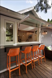 Outdoor bar with a sliding window, four bright orange bar stools, and a wooden deck.