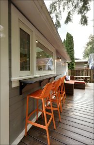 Outdoor patio with a high counter and three orange bar stools. Adjacent to a house with a sliding window and wooden deck flooring, surrounded by trees and fences.