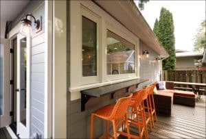 Outdoor patio with orange bar stools at a counter under a window, a wicker sofa set on wooden decking, and a lit wall lamp.