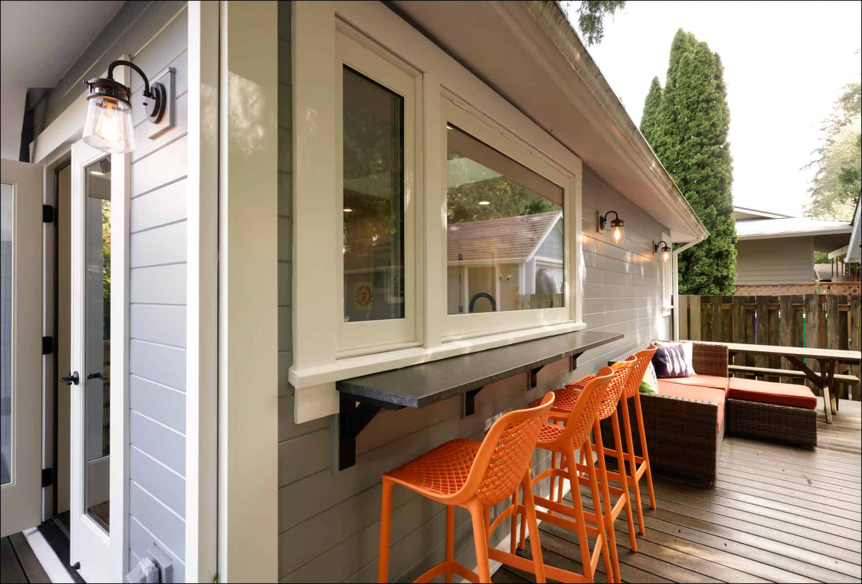 Outdoor patio with orange bar stools at a counter under a window, a wicker sofa set on wooden decking, and a lit wall lamp. Outdoor patio with orange bar stools at a counter under a window, a wicker sofa set on wooden decking, and a lit wall lamp.