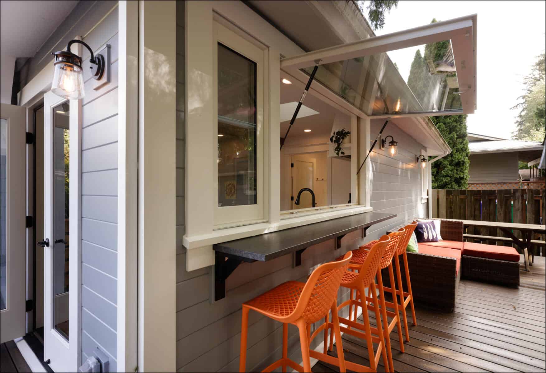 Outdoor kitchen area with a serving window, four orange bar stools, and a wooden deck featuring outdoor seating and a fence. Outdoor kitchen area with a serving window, four orange bar stools, and a wooden deck featuring outdoor seating and a fence.
