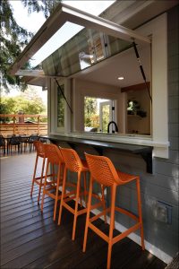 Outdoor bar area with a large window opening to the kitchen, featuring three orange barstools on a wooden deck.