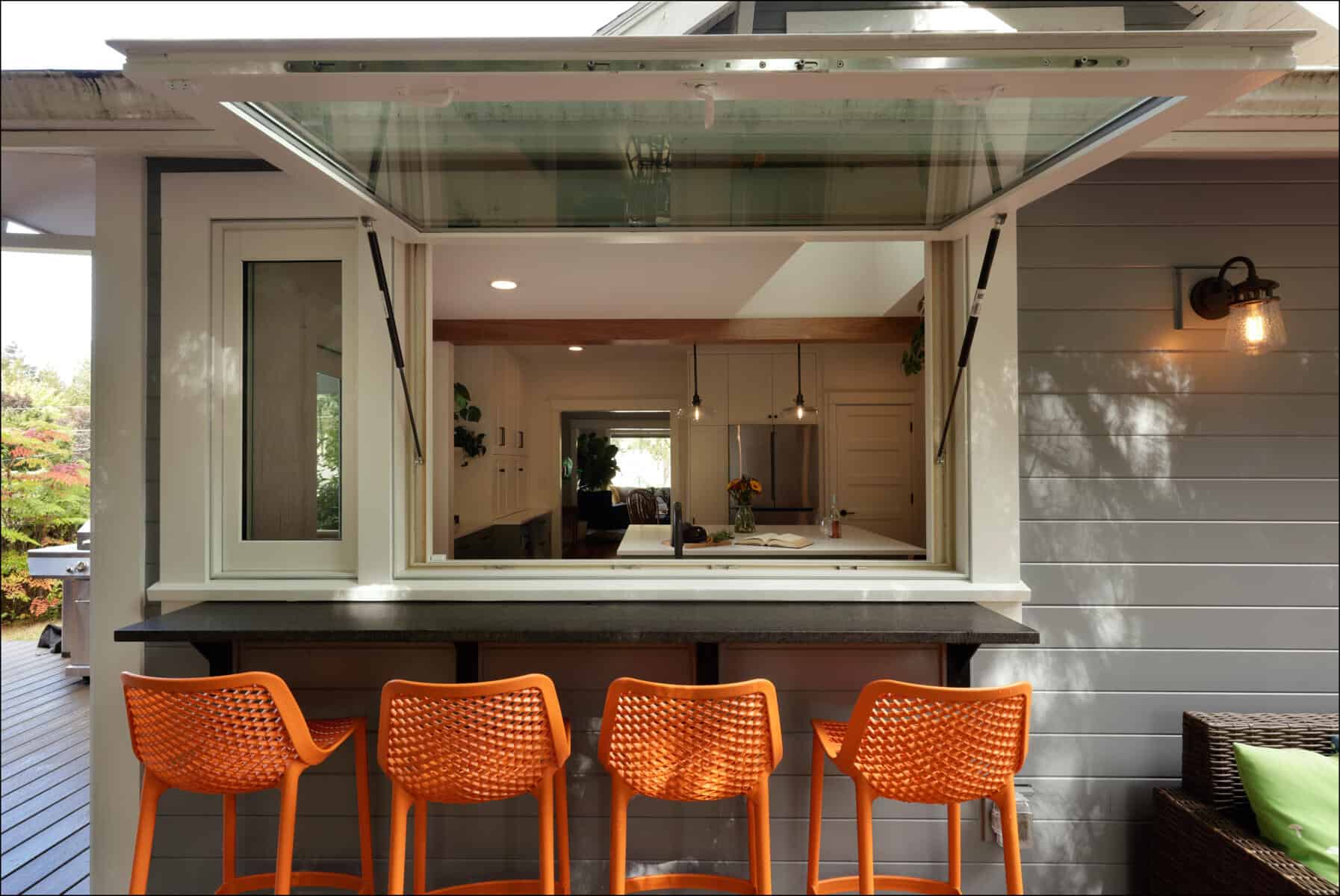 A kitchen with an open window hatch looks out onto a patio with four orange chairs lined up along a countertop. A kitchen with an open window hatch looks out onto a patio with four orange chairs lined up along a countertop.