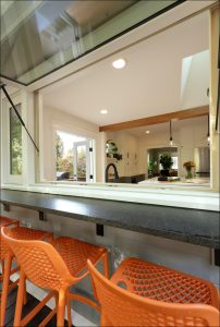 Kitchen with a pass-through window opening to a counter with three orange stools. Indoor view shows white cabinets and a sink, and outdoor greenery is visible through the window.