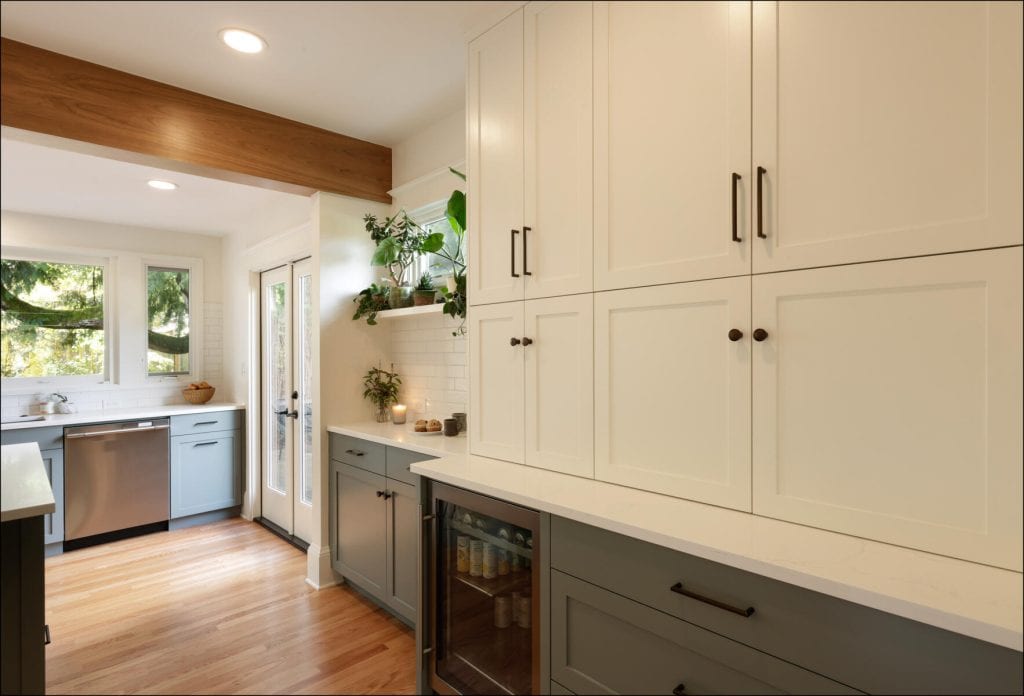 A modern kitchen with white cabinetry, wooden flooring, and stainless steel appliances. Large windows provide natural light. Green plants add a touch of color.