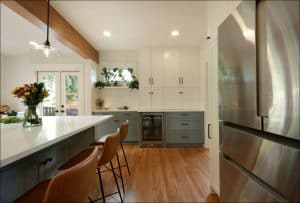 Modern kitchen with white and grey cabinets, wooden floor, a large island with leather bar stools, a stainless steel fridge, and a glass door leading outside.