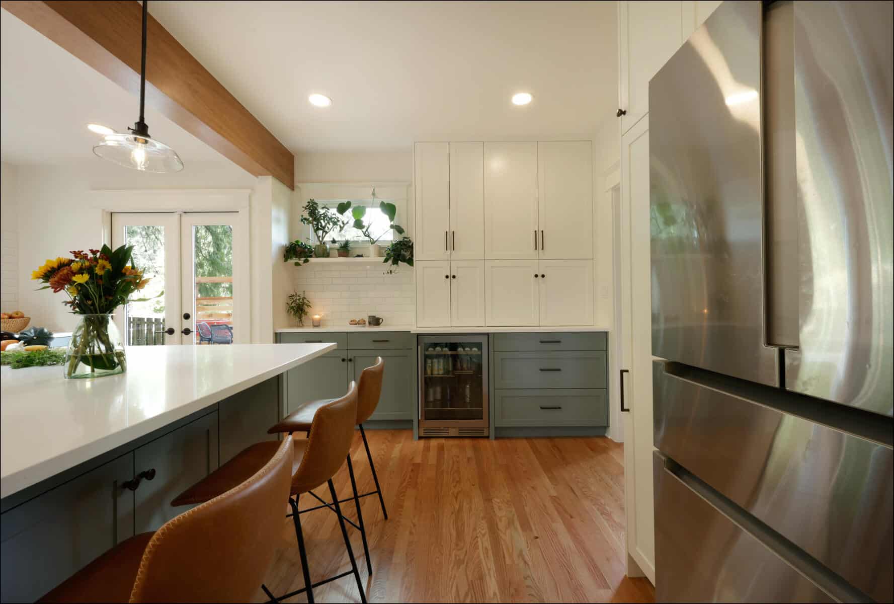 Modern kitchen with white and grey cabinets, wooden floor, a large island with leather bar stools, a stainless steel fridge, and a glass door leading outside. Modern kitchen with white and grey cabinets, wooden floor, a large island with leather bar stools, a stainless steel fridge, and a glass door leading outside.
