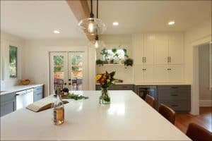 Modern kitchen with a large island featuring a vase of flowers, a book, and a bottle. White cabinets and grey drawers are in the background. Two pendant lights hang above the island.