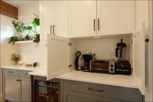 A kitchen counter with white cabinets, open to reveal a coffee maker, toaster, and blender. Nearby, there is a small fridge, potted plants, and a window with natural light.