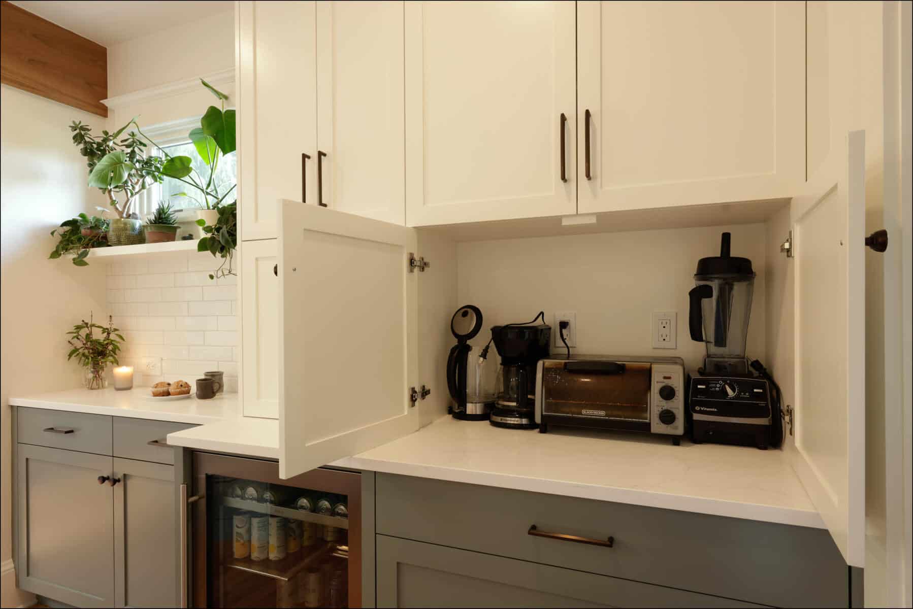 A kitchen counter with white cabinets, open to reveal a coffee maker, toaster, and blender. Nearby, there is a small fridge, potted plants, and a window with natural light. A kitchen counter with white cabinets, open to reveal a coffee maker, toaster, and blender. Nearby, there is a small fridge, potted plants, and a window with natural light.