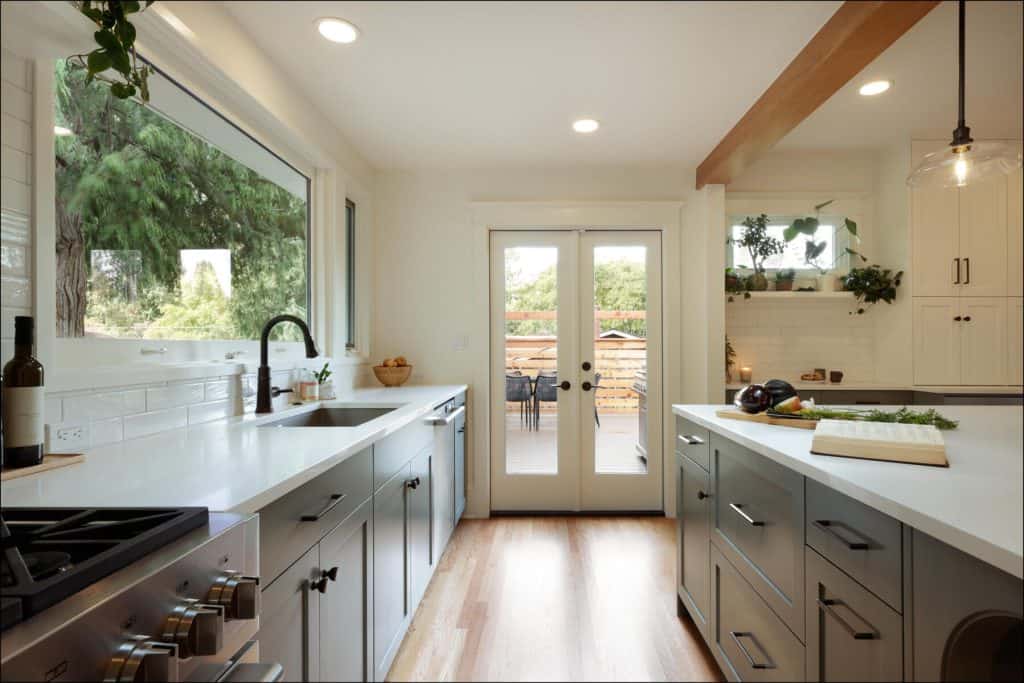 Modern kitchen with white cabinets, large windows, and wooden accents. Double doors lead to a patio with outdoor seating. Kitchen island has a chopping board with herbs and vegetables.