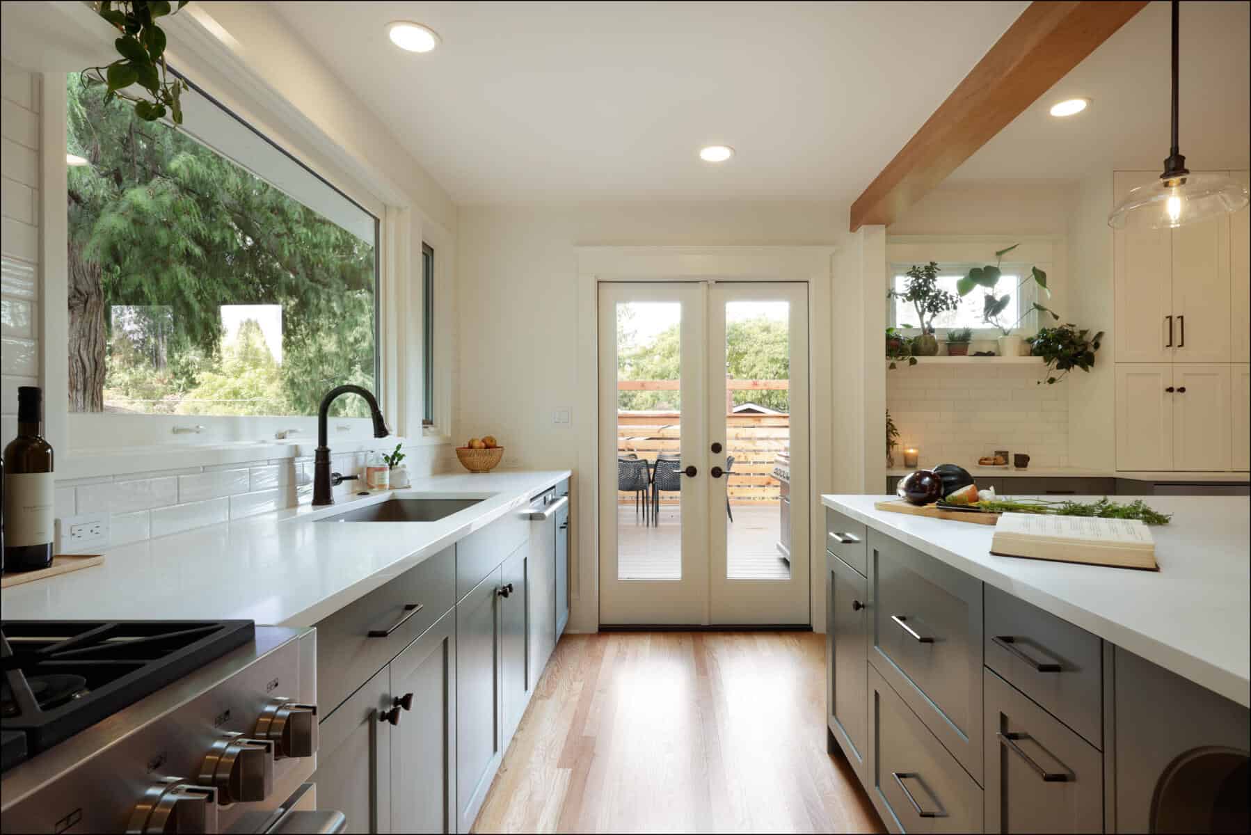 Modern kitchen with white cabinets, large windows, and wooden accents. Double doors lead to a patio with outdoor seating. Kitchen island has a chopping board with herbs and vegetables. Modern kitchen with white cabinets, large windows, and wooden accents. Double doors lead to a patio with outdoor seating. Kitchen island has a chopping board with herbs and vegetables.