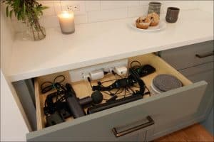 Drawer filled with electronic devices, cords, two round pads, and a massage gun. Marble countertop above holds a candle, two muffins on a plate, and two mugs.