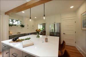 Modern kitchen with white cabinets, stainless steel fridge, and an island with a cookbook, flowers, and vegetables. Two pendant lights hang above the island.
