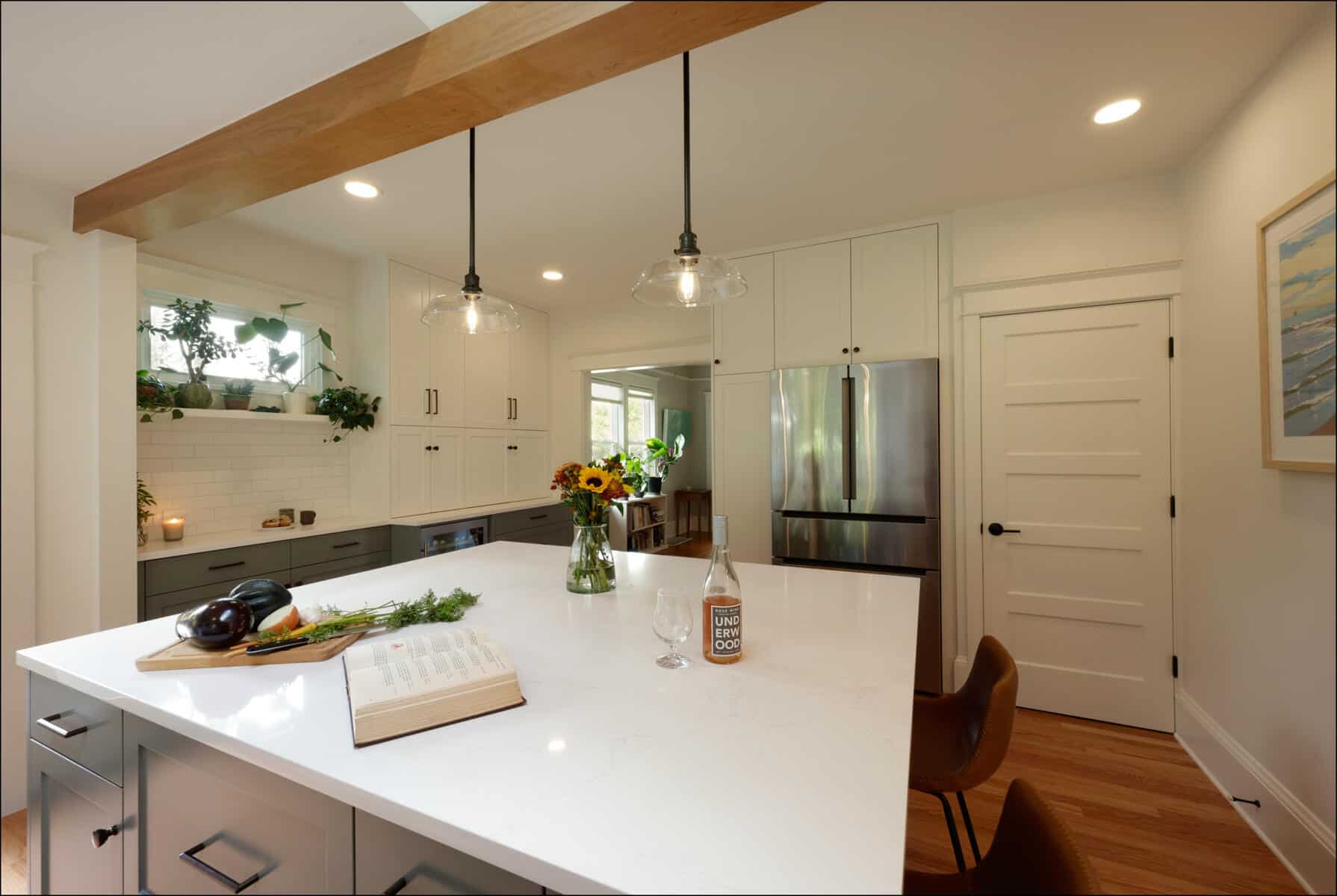 Modern kitchen with white cabinets, stainless steel fridge, and an island with a cookbook, flowers, and vegetables. Two pendant lights hang above the island. Modern kitchen with white cabinets, stainless steel fridge, and an island with a cookbook, flowers, and vegetables. Two pendant lights hang above the island.