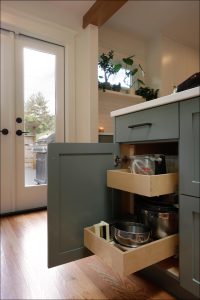 A kitchen cabinet with open pull-out drawers revealing kitchenware and utensils, set in a bright kitchen with large glass doors and potted plants on a shelf.