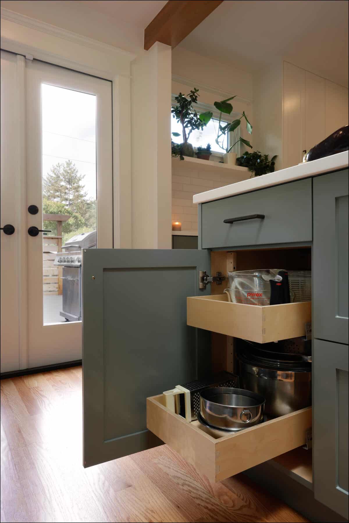 A kitchen cabinet with open pull-out drawers revealing kitchenware and utensils, set in a bright kitchen with large glass doors and potted plants on a shelf. A kitchen cabinet with open pull-out drawers revealing kitchenware and utensils, set in a bright kitchen with large glass doors and potted plants on a shelf.