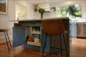 Kitchen with a blue island, open cabinet showing pantry items, two brown bar stools, a vase with flowers, and a large window with a view of trees.