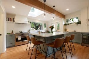 Modern kitchen with a large island, four brown chairs, stainless steel appliances, and overhead lights. Skylight and windows provide natural light, with plants and books on the shelves.