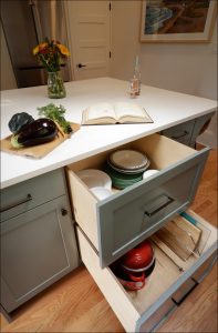 A kitchen island with open drawers displaying plates and cookware, a countertop with an open cookbook, vegetables, a vase with flowers, and a bottle of wine.
