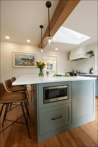Modern kitchen with a large island featuring a built-in microwave, overhead lights, and wooden elements. There are bar stools, a vase of flowers, and vegetables on the counter.