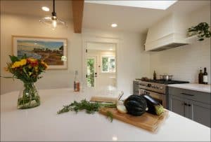A modern kitchen with a white countertop, vegetables on a cutting board, a vase of flowers, and a stainless steel stove. A large window lets in natural light.