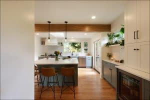 Modern kitchen with a wooden floor, island with brown chairs, pendant lights, white cabinets, green plants, and natural light from large windows and a glass door.