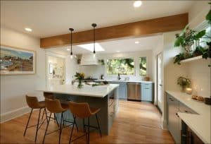 Modern kitchen with a central island, three barstools, pendant lights, and plants. White walls, wooden floors, and large windows create a bright atmosphere.