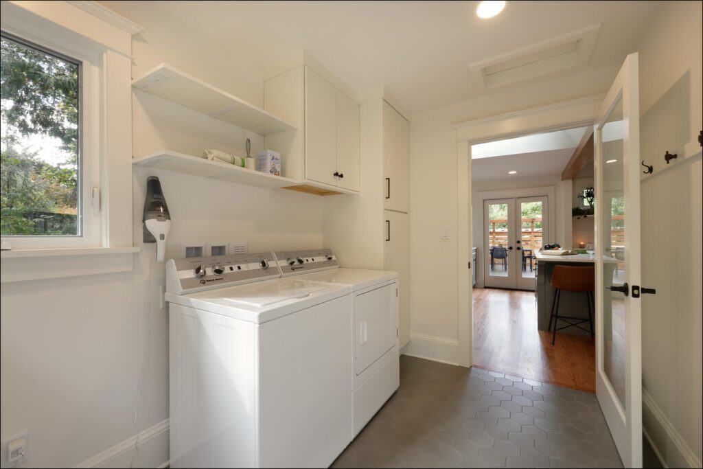 A bright laundry room with a window, white washer and dryer, shelf with cleaning supplies, and an open door leading to a kitchen area.