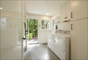 A bright laundry room with a washer and dryer, white cabinets, and a glass door leading outside. The floor has hexagonal tiles, and a mat is placed by the door.