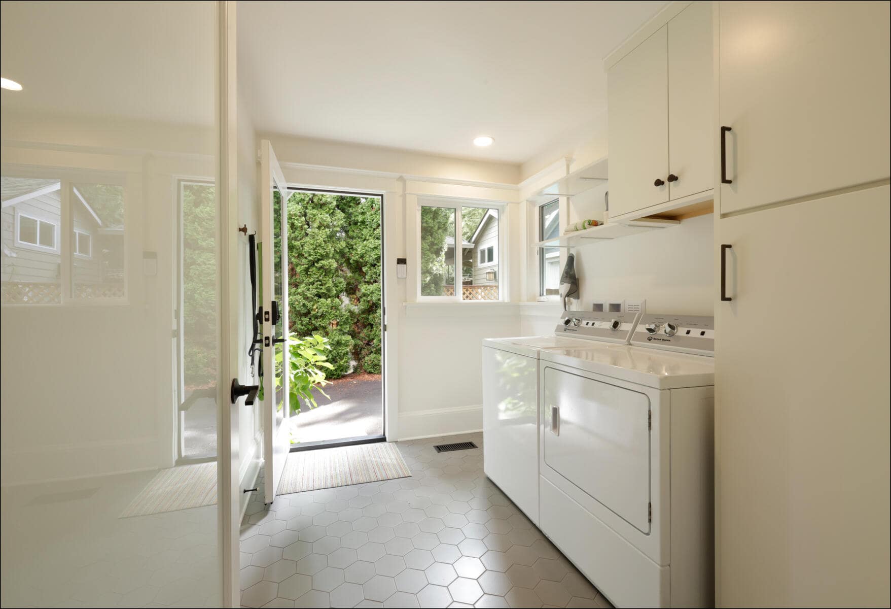 A bright laundry room with a washer and dryer, white cabinets, and a glass door leading outside. The floor has hexagonal tiles, and a mat is placed by the door. A bright laundry room with a washer and dryer, white cabinets, and a glass door leading outside. The floor has hexagonal tiles, and a mat is placed by the door.