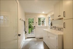 A laundry room with a washer and dryer, white cabinets, and a door leading outside. Natural light enters through the windows, illuminating hexagonal tile flooring.