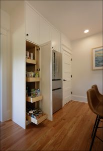 A modern kitchen with a tall pantry cabinet slightly open, revealing shelves stocked with various food items. Adjacent is a stainless steel refrigerator and two brown leather chairs.