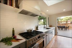 Modern kitchen with stainless steel stove, white subway tile backsplash, and wooden countertops. Fresh vegetables and olive oil bottle on the counter. Opens to a deck with outdoor seating.