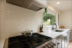Modern kitchen with a black stove, steel pot, white countertops, sink, and large windows. White subway tiles on the wall and potted plants near the sink.