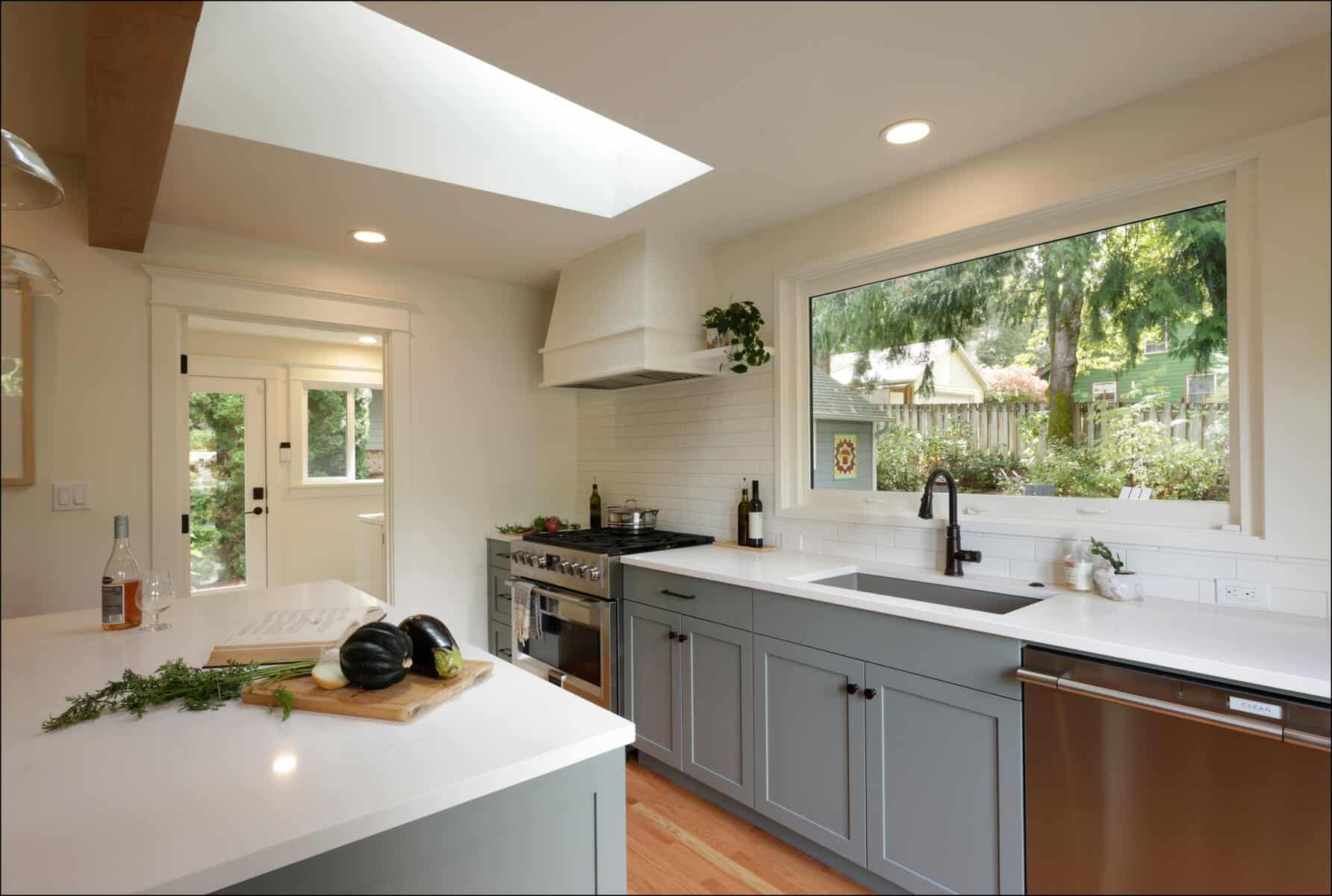 A modern kitchen with light gray cabinets, a white countertop holding vegetables and herbs, a gas stove with a hood, and a large window overlooking a garden. A modern kitchen with light gray cabinets, a white countertop holding vegetables and herbs, a gas stove with a hood, and a large window overlooking a garden.