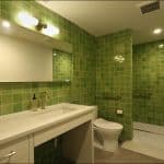 Bathroom with green tiles, a large mirror, sink with toiletries, toilet, and a walk-in shower featuring a bench and towel rack&mdash;perfectly showcasing home remodeling elegance.